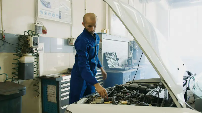Majstor u plavom kombinezonu pregleda motor bijelog auta ispod otvorene haube